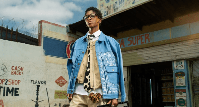 A person with defiant optimism, wearing layered clothing and glasses, stands outside a shop with painted signs, holding a small bag.