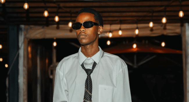 A person with a creative bag, wearing sunglasses, a light-colored shirt, and a patterned tie stands under hanging string lights at night.