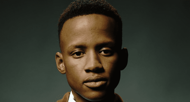 A young man with short hair looks directly at the camera, wearing a brown jacket and white shirt, against a plain dark background—artist Kane Keid from debut album SON OF SOiL.