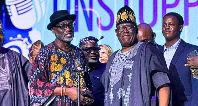 A group of men in traditional attire stand on stage at the AFRIMA 2025, two shaking hands as Kenny Ogungbe receives one of the Lifetime Achievement Awards, with a microphone and audience visible in the background.