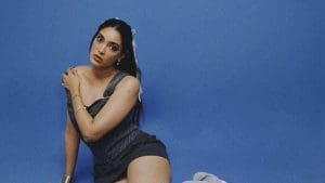Indian singer Khevna sits on the floor in a dark sleeveless outfit against a plain blue background, looking at the camera with a neutral expression, embodying an Afro-Pop vibe.