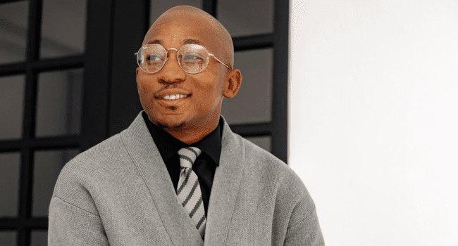A person wearing glasses, a grey cardigan, a black shirt, and a striped tie stands in front of a window and white wall, looking to the side and smiling in what feels like a Tribute To AKA on his Heavenly Birthday.