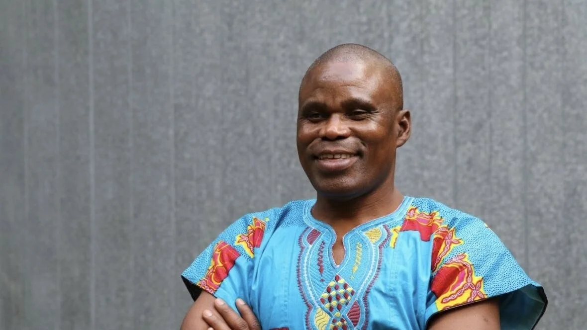 A man in a blue patterned traditional shirt stands with his arms crossed, smiling in front of a plain gray background, capturing the spirit of Celebrate Africa and Ghanaian music at CapU.