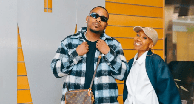 Two people pose in front of an orange and gray wall, channeling new season style; one wears sunglasses, a plaid jacket, and a crossbody bag, while the other sports a cap, hoop earrings, and a white shirt.