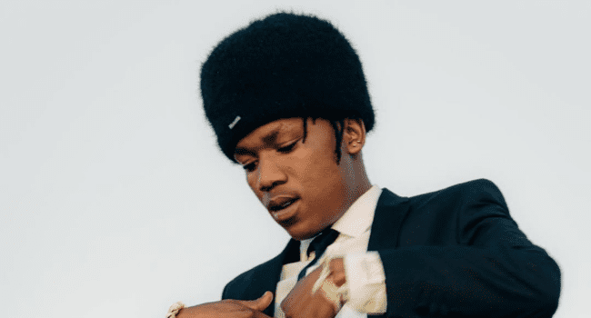 A man wearing a dark suit and a black fuzzy hat looks down and adjusts his clothing against a plain light background, hinting at the poised style featured on the MR BULLY album by LaCabra.