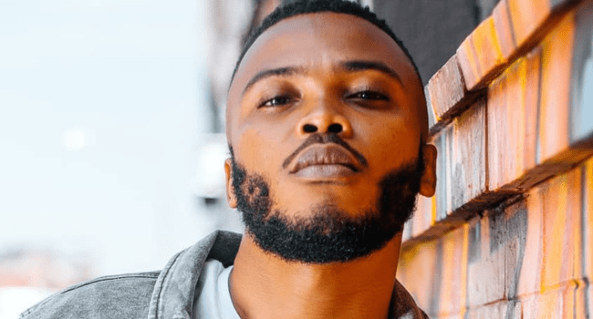 A man with a short beard and mustache stands outdoors, leaning against a brick wall and looking directly at the camera, wearing a Summer Ties shirt.