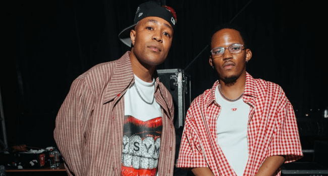 Two men stand side by side at the Milk + Cookies Festival, both wearing red and white shirts; one wears a cap backwards and a graphic tee, the other has glasses and a checkered shirt—perhaps ready for surprise cameos.