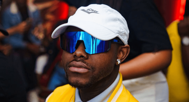 A man wearing a white cap, reflective blue sunglasses, diamond earrings, and a yellow jacket sits indoors with a blurred crowd in the background, deep in thought about his next album and the creative process behind it.
