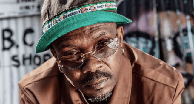 A man wearing glasses, a green bucket hat, and a brown jacket looks directly at the camera; graffiti is visible in the background, reflecting the vibrant energy often seen on digital platforms.