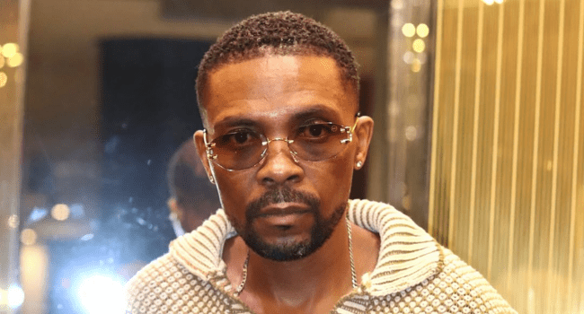 A man wearing glasses and a striped sweater looks directly at the camera, standing indoors with a softly lit background, reminiscent of Sizwe Dhlomo’s confident presence.