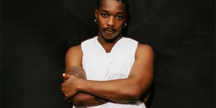 A person with braided hair and a mustache wears a sleeveless white top, standing with arms crossed against a dark background, embodying an Everyday style reminiscent of Magixx in 2026.