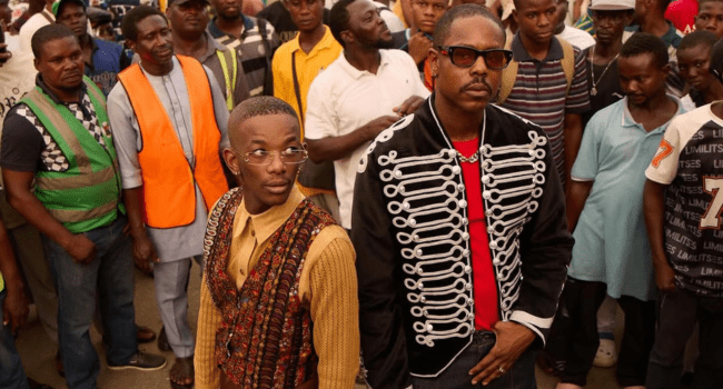 Two men in distinctive outfits stand in front of a crowd outdoors. One, channeling Motho Waka style with a patterned vest, stands beside another in a black jacket with white trim and sunglasses.