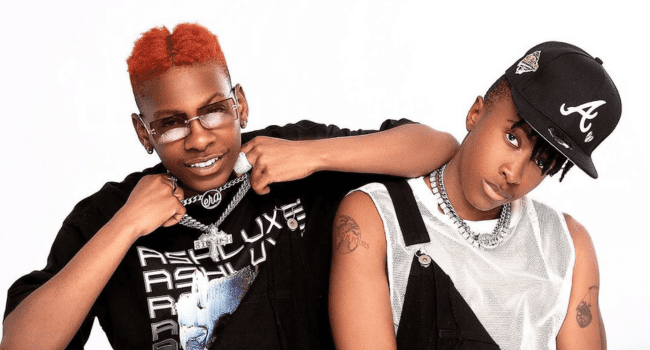 Two people pose together in studio mode against a white background; one wears a black shirt, orange hair, and glasses, while the other sports a cap, overalls, and a sleeveless shirt.