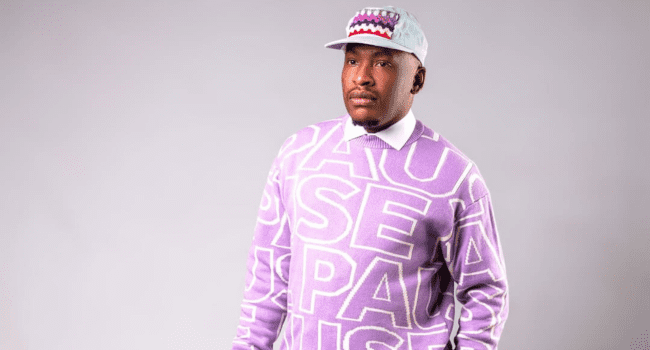 During a 2026 studio session, a person wearing a pale blue cap and a lavender sweater with large white text stands against a plain light gray background.