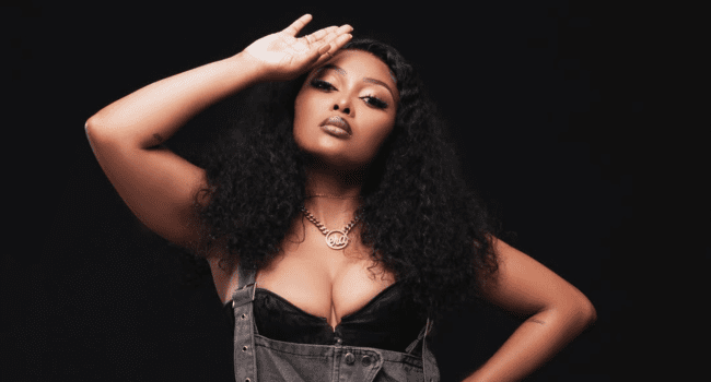 A woman with long curly hair poses with one hand raised to her forehead, wearing a black top, denim overalls, and a necklace, against a dark background—radiating confidence like Miss Pru in the face of body shamers.