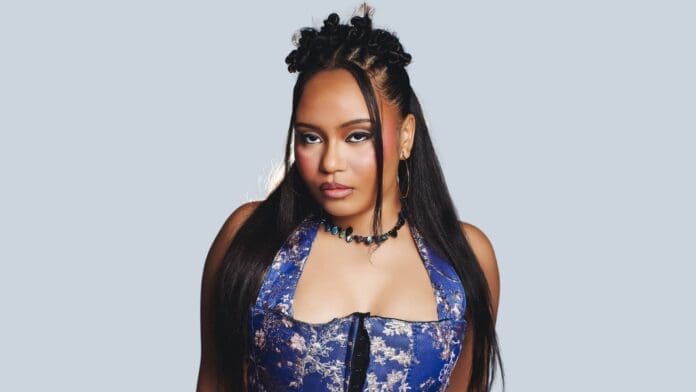 A woman with long straight hair and styled buns, reminiscent of Spotify’s Equal artist Moliy, wears a blue patterned halter top and a beaded necklace, posing against a plain light background.