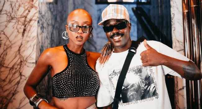 Two people pose together indoors; one wears a black studded crop top and large hoop earrings, while Uncle PartyTime sports sunglasses, a plaid cap, and a graphic t-shirt, giving a thumbs up at this lively birthday tribute.
