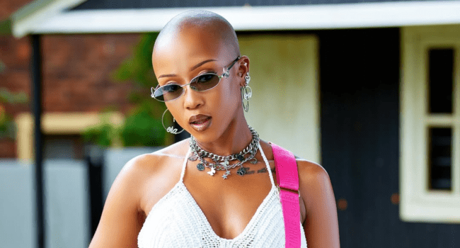 Moozlie, with a shaved head, sunglasses, layered necklaces, earrings, and a white crochet top, stands outdoors in front of a black and white building.