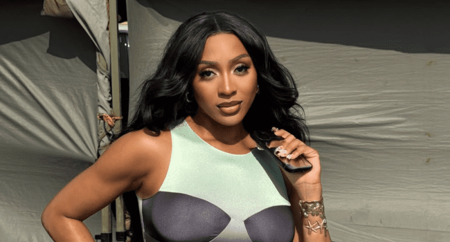 A woman with long black hair poses outdoors, wearing a form-fitting sleeveless dress and holding a pair of sunglasses in her hand, capturing the confident style often seen on Nadia Nakai.