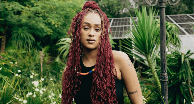 Person with long, burgundy dreadlocks and a black sleeveless top stands outdoors among lush green plants and a glass structure, embodying true artistry in an AI era.