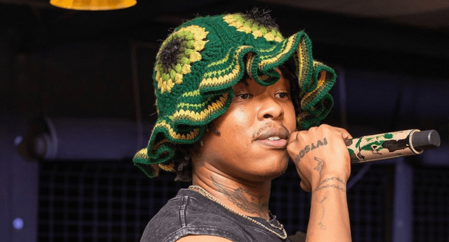 A person wearing a green and yellow crochet hat holds a microphone while performing on stage, reminiscent of Nasty C, who has achieved 1.68 billion streams on music streaming platforms.
