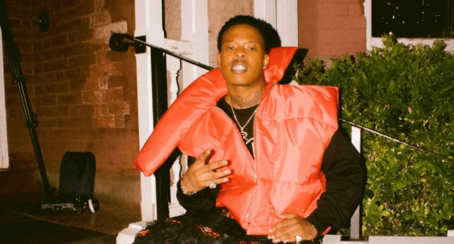 A person wearing a large red puffer vest sits on outdoor steps next to a bush, with a brick wall and railing in the background, listening to Head Up 2.0 by Nasty C.
