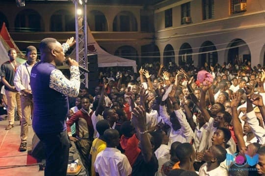 A man on stage speaks into a microphone to a large, enthusiastic crowd at an outdoor High School Praise concert at night; many audience members have their hands raised, enjoying the lively atmosphere and an energetic Amapiano song.