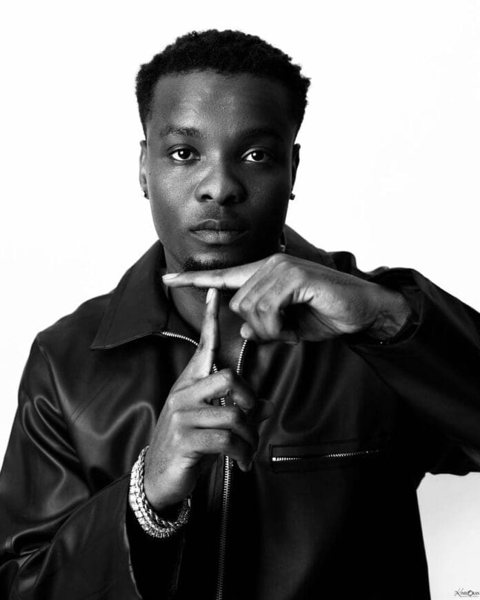 Black and white portrait of a man in a leather jacket making a “time out” gesture with his hands, channeling Lagos style and Afrobeats energy as he looks directly at the camera.