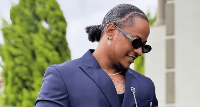A man in a dark blue suit with sunglasses and earrings stands outdoors, looking down and smiling—a style reminiscent of Priddy Ugly in the "Bags on Bags" era.