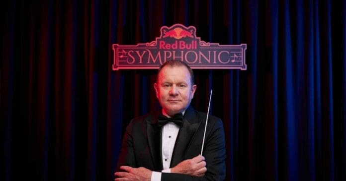 A man in a tuxedo, possibly famed Sowetan conductor Adam Howard, holds a baton while standing before a "Red Bull Symphonic" sign with a dark curtain backdrop, ready to blend classical music with Amapiano rhythms.