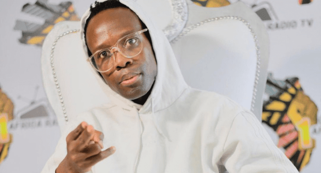 A person wearing glasses and a white hoodie sits on a white chair, gesturing with their hand, with a logo-filled backdrop behind them at the Timeline Radio Awards, celebrating Hip-Hop Legends.