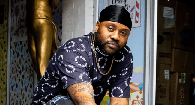 A man wearing a black durag, printed t-shirt, and gold chain sits in front of a store with colorful posters and boxes—perhaps reflecting on his Dormant Music Relationship or channeling the cool vibe of Stogie T.