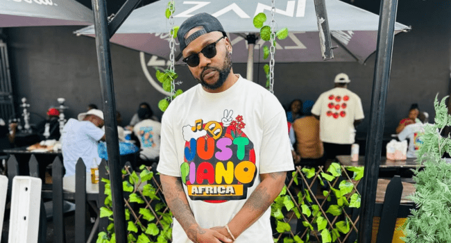 A man wearing sunglasses, a cap, and a "Piano Africa" t-shirt stands outdoors in front of a restaurant patio, where people are seated in the background—channeling vibes reminiscent of Cassper's cool style.