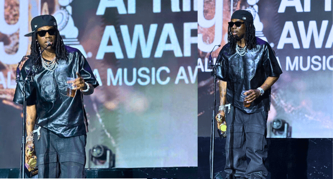 A man in sunglasses, black shirt, and baggy pants stands on stage holding a drink and a microphone, with "AWARD" visible on the backdrop behind him at the AFRIMA ceremony.