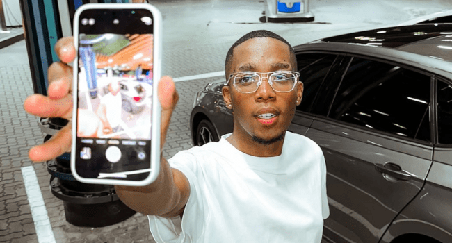 A person wearing glasses and a white shirt holds a smartphone up for a Freestyle Fridays selfie at a gas station next to a dark-colored car at night, capturing the Roiii vibe.