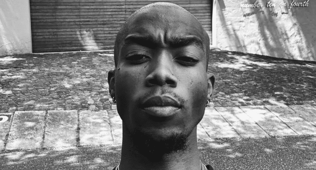 A person with a neutral expression stands outside on a paved street, with a garage door and white building in the background. The black and white image has an R&B vibe, reminiscent of an old album cover or vintage tape.