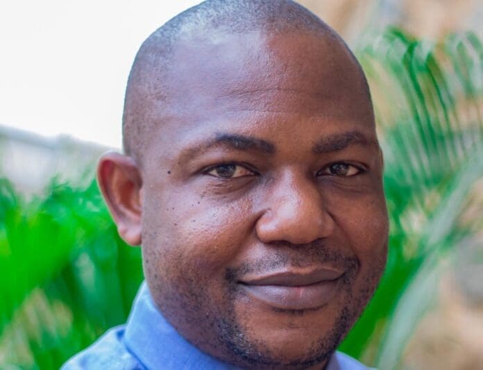 A man with short hair and a trimmed beard, wearing a blue collared shirt, stands outdoors with greenery in the background, reflecting on ways to rebuild music education within the African education system.