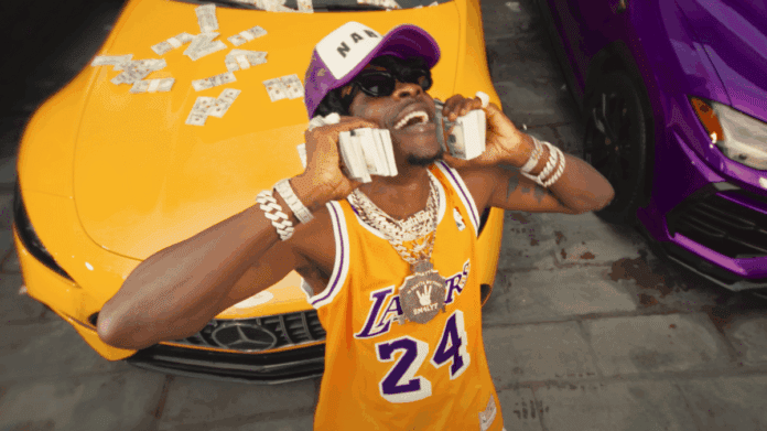 A man wearing a Lakers jersey, cap, and jewelry holds stacks of cash to his ears next to a yellow car with more cash on its hood, showing off the swagger of "A Taste Of The African King," inspired by Shatta Wale's new project.