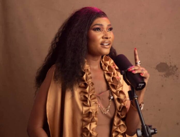 A woman with long wavy hair speaks into a microphone, wearing a beige outfit with ruffled details. Channeling Afro Pop vibes, she stands confidently against a plain background, reminiscent of an Eleas Modelà performance.