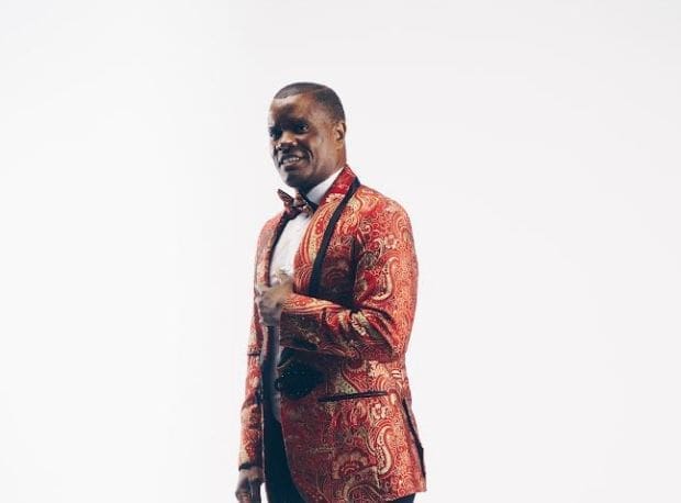 A man stands against a plain white background wearing a red paisley-patterned suit jacket, white shirt, and bow tie, embodying the vibrant style often seen in gospel music artists like Mfumu ya Mafumu Ngumuya.