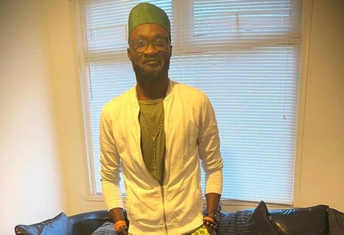 A man wearing a green cap, glasses, a white jacket, and olive green pants stands indoors in front of a window with blinds, channeling Taiwo Akintoye’s signature style inspired by African Tech trends.