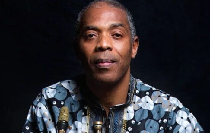 A man with short gray hair wearing a patterned shirt sits in front of a dark background, with two saxophones visible—a scene echoing the musical legacy of Femi Kuti and the enduring spirit of Nigeria's challenges.