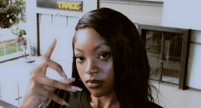 A woman with long black hair and decorated nails poses, holding her hand up near her face, standing outside a building with a "TRADE" sign in the background, showcasing the 066 Deluxe Release from Scumie.