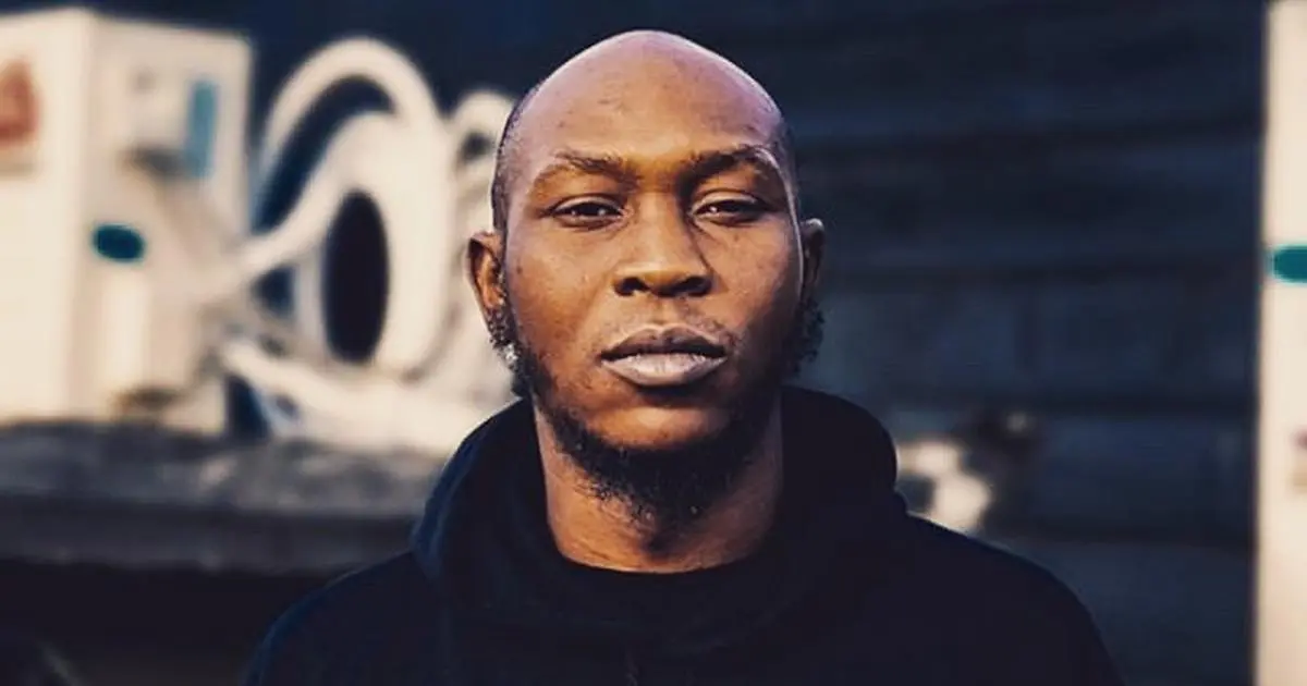 A man with a shaved head and facial hair looks directly at the camera, wearing a black hoodie. The background is blurred with industrial equipment visible, evoking the bold style of Afrobeats artist Seun Kuti.