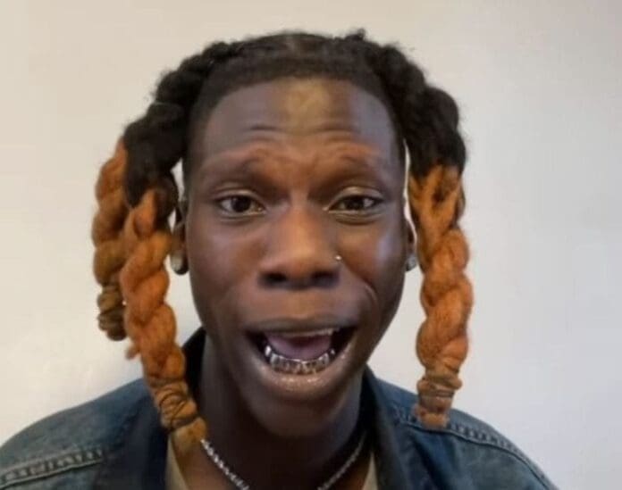 A person with orange-tipped, twisted braids, a nose piercing, and visible dental grills poses against a plain background—a look inspired by the Neo-fuji style popularized by Seyi Vibez, featured in The Guardian Nigeria.