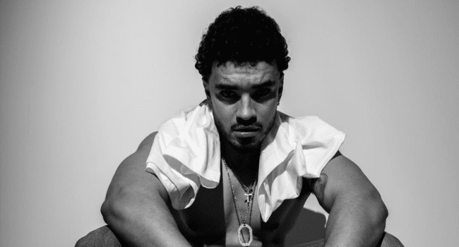 A man with curly hair sits shirtless, an Afro Comb nearby, wearing a cross necklace and an open white shirt, looking directly at the camera with a serious expression against a plain background, reminiscent of a Shane Eagle music video.