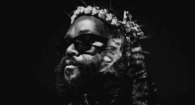 A person with a beard and sunglasses wears a decorative headpiece with feathers and flowers, photographed in black and white against a dark background—evoking the striking style of Sjava’s forthcoming album, INkanyezi NeZinkanyezi.