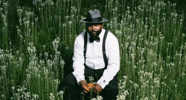 A man wearing a white shirt, black suspenders, bow tie, and gray hat sits in a field of tall white wildflowers, listening to Sjava’s INKANYEZI NEZINKANYEZI on Apple Music as he looks at the camera.
