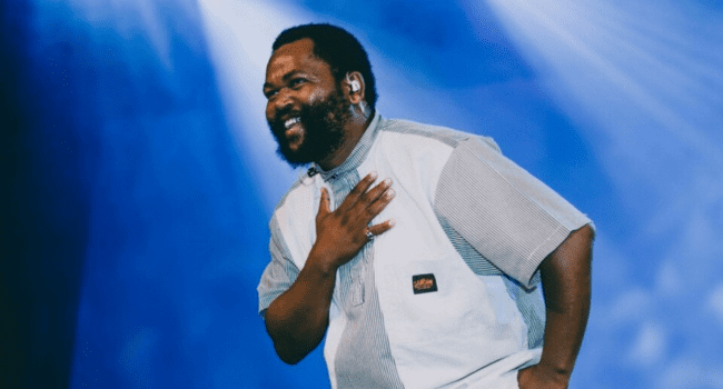 A man with a beard and short hair, resembling Sjava, smiles and places his hand on his chest while standing under blue stage lighting—capturing the spirit of genre confusion celebrated by listeners on Spotify.