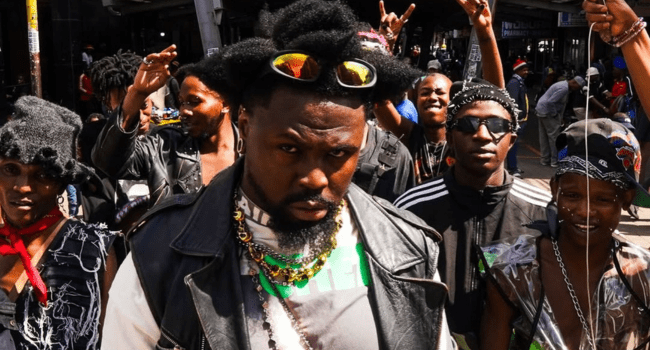 A group of people dressed in bold, eclectic streetwear pose outdoors, channeling music video vibes, with one man in front—reminiscent of Stilo Magolide—sporting sunglasses on his head and layered necklaces.